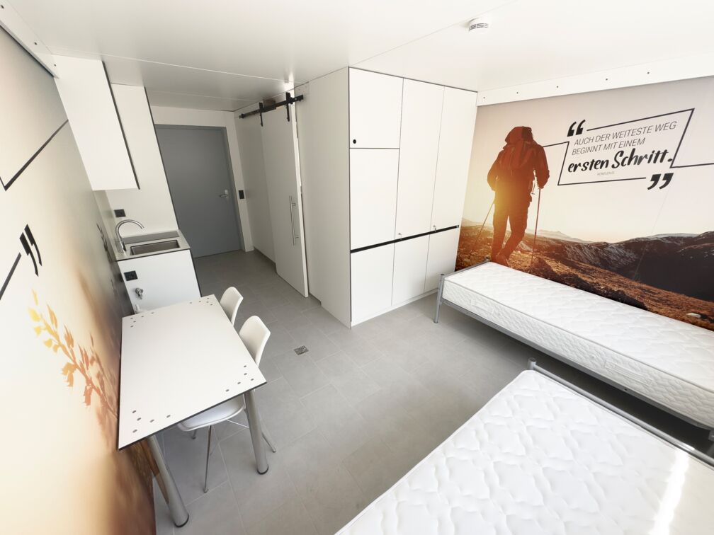 Helles Zimmer mit zwei Betten, Schreibtisch, Schrank und Wandbild.